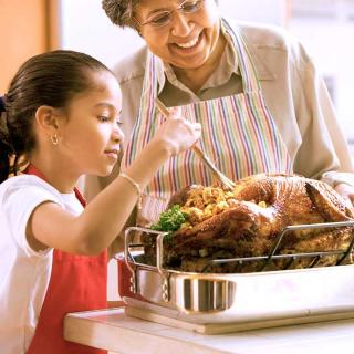 A young girl bastes a turkey in the kitchen while her grandmother watches.